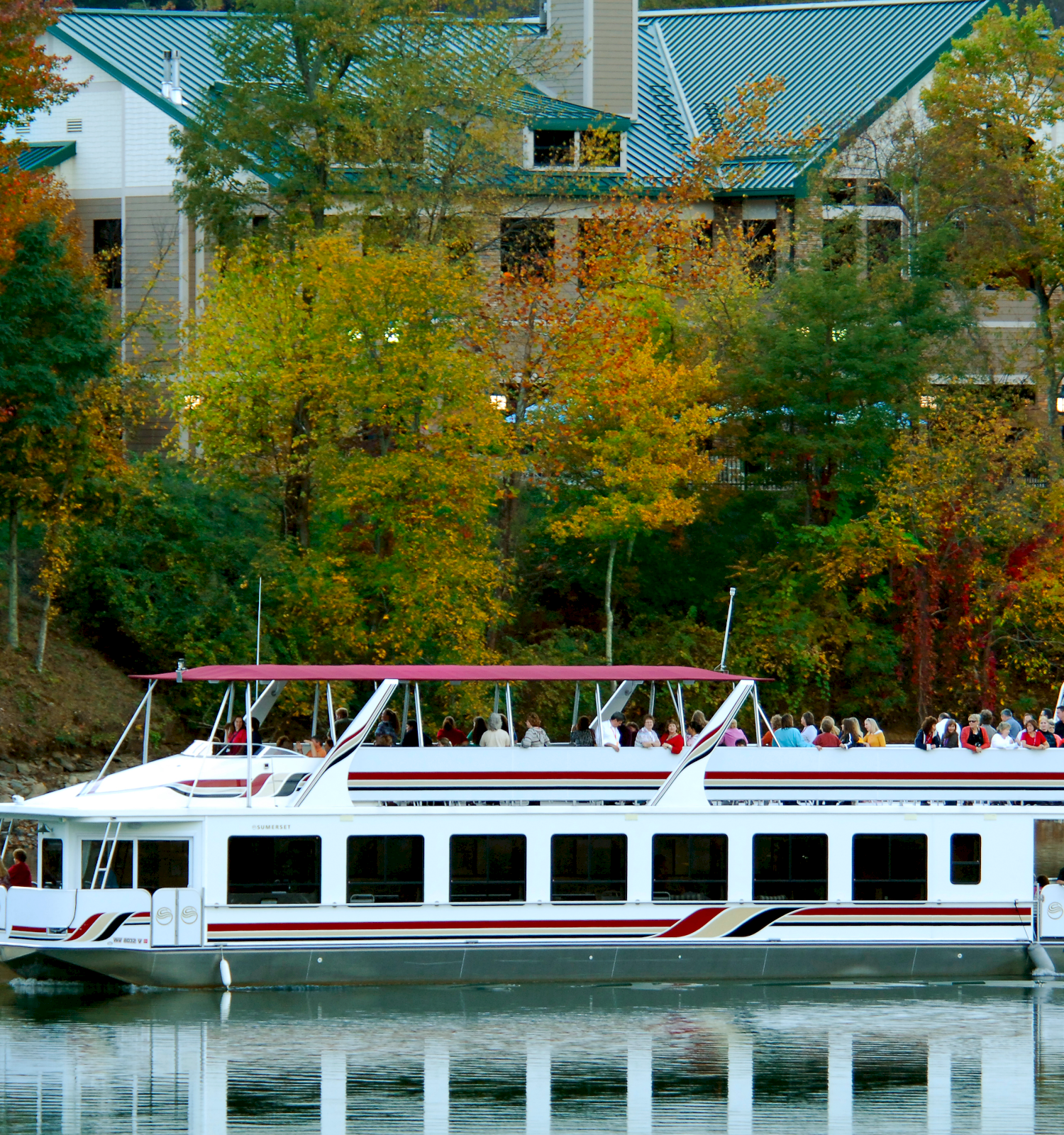 A white double-deck boat sails on a calm lake, surrounded by autumn trees and a building in the background.