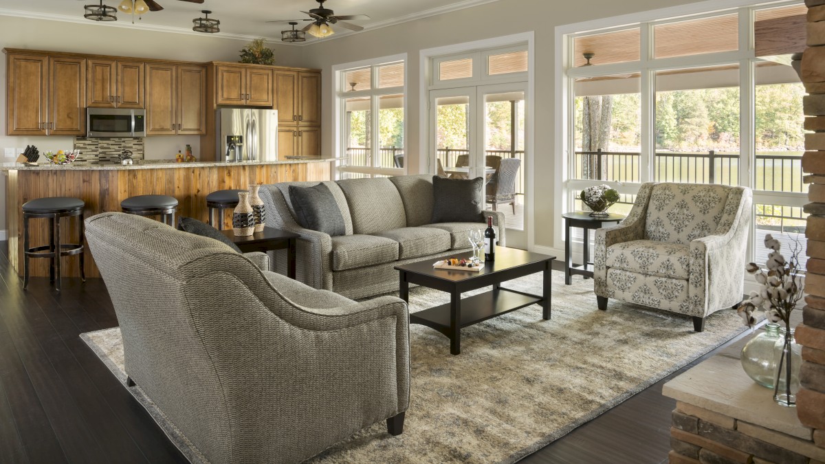 This image shows a modern living room with a grey couch and armchairs, a rug, a coffee table, and a kitchen area with bar stools and wooden cabinets.