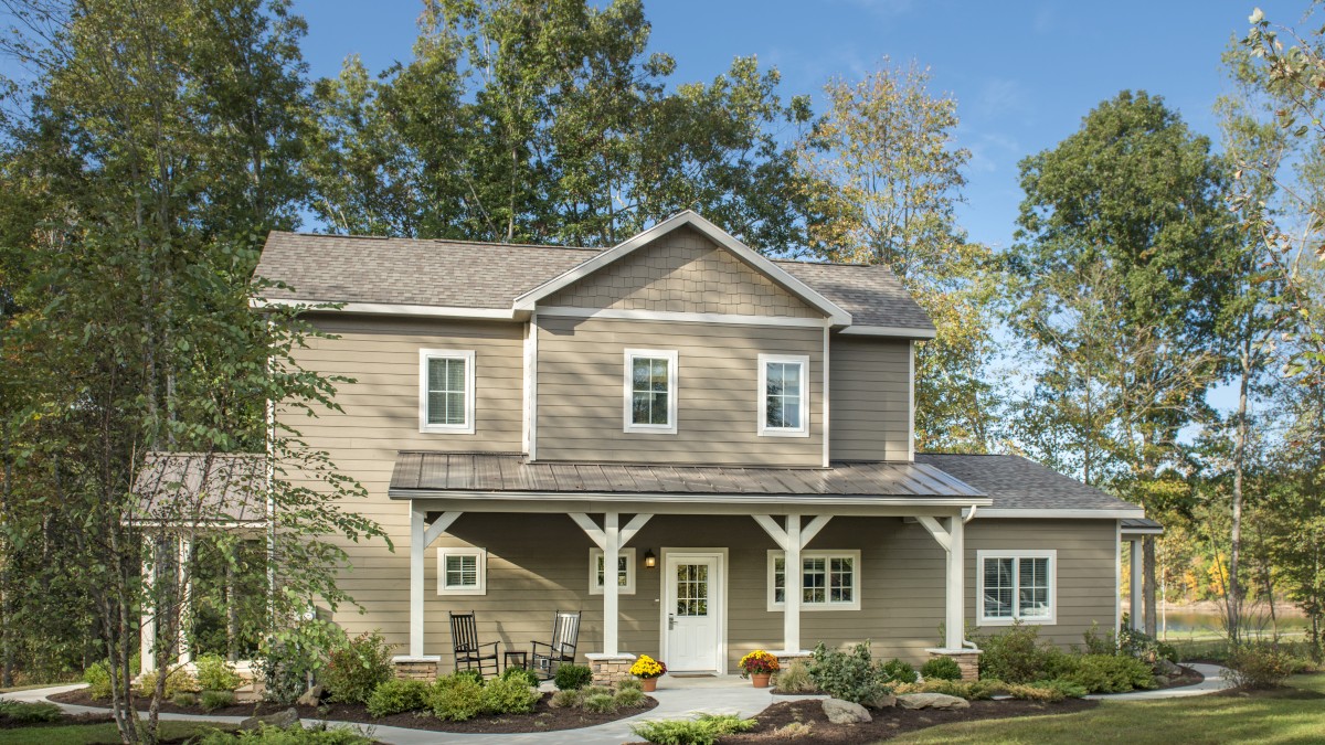 A two-story house with beige siding, a covered front porch, and a landscaped yard surrounded by trees under a clear blue sky.