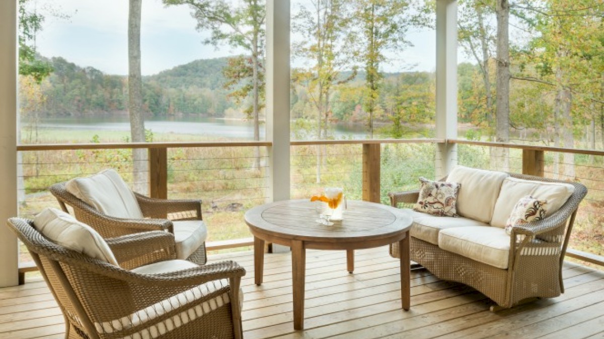 A cozy porch with wicker furniture, including a sofa and two chairs around a round wooden table, overlooking a scenic lake and forest view.