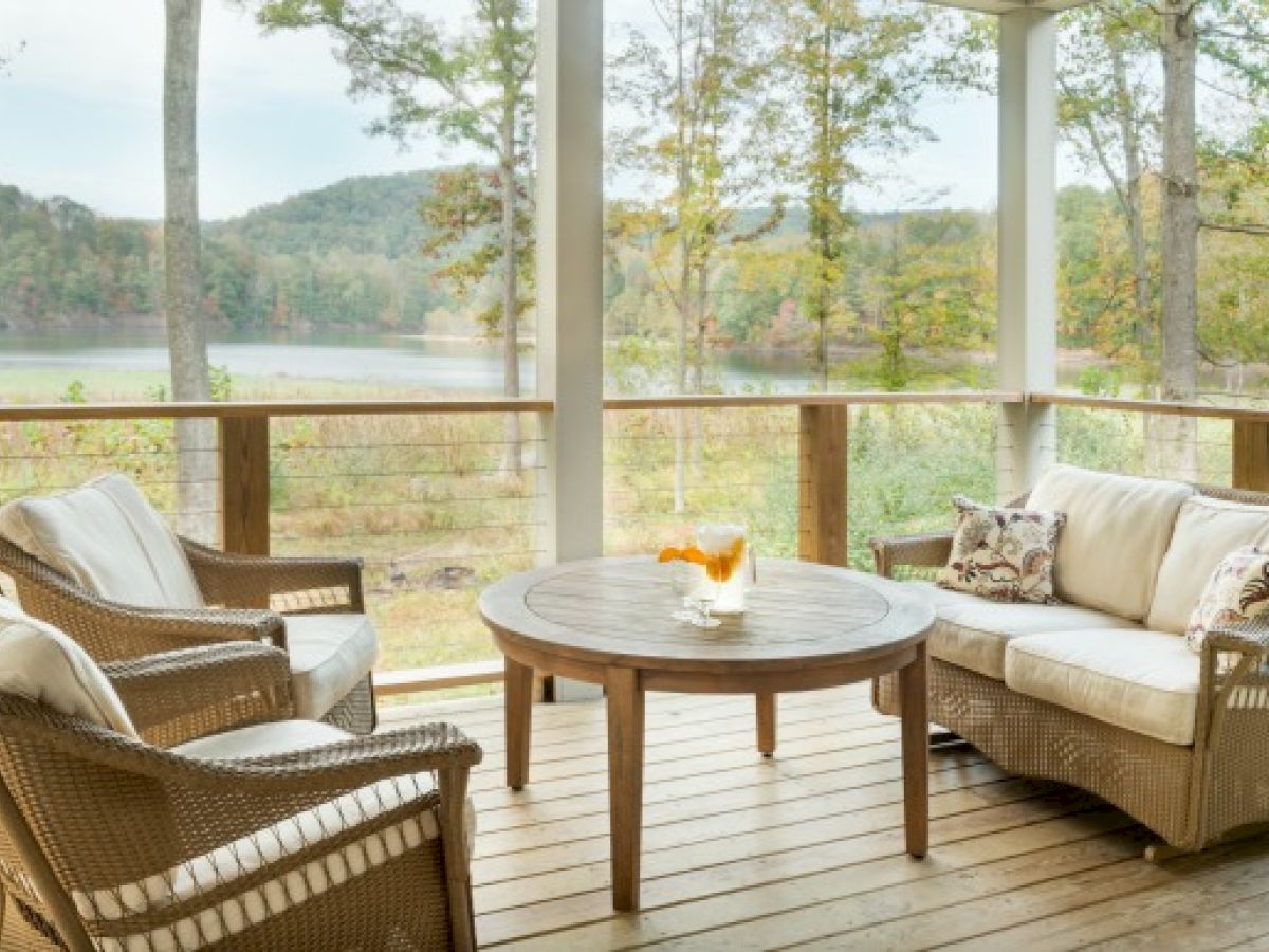 A cozy porch with wicker furniture, including a sofa and two chairs around a round wooden table, overlooking a scenic lake and forest view.