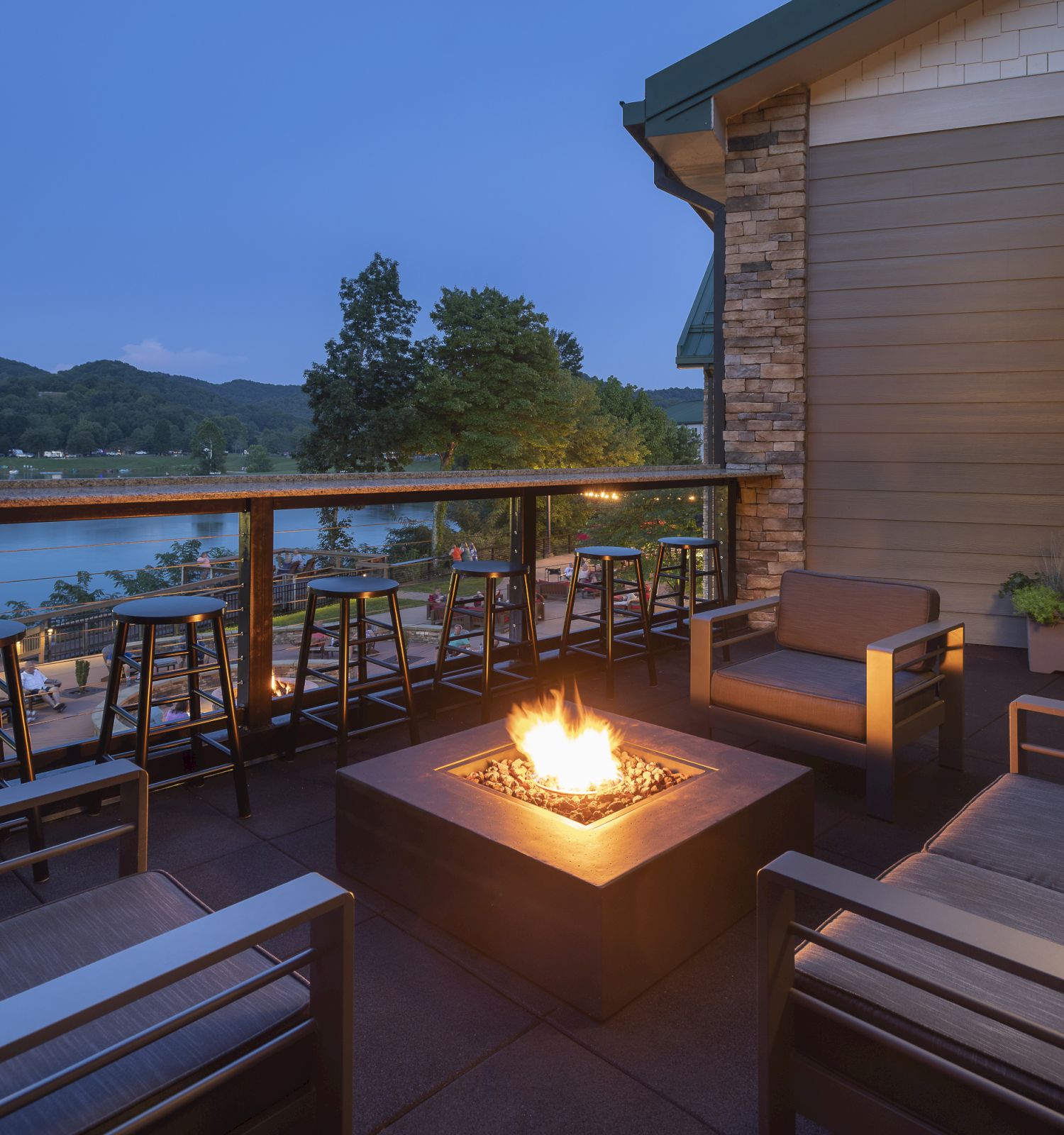An outdoor patio with a fire pit, surrounded by chairs and a railing with barstools, overlooking a scenic lake and mountains in the evening.