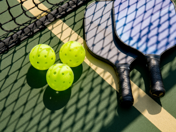 The image shows two pickleball paddles and three yellow pickleball balls on a court near the net.