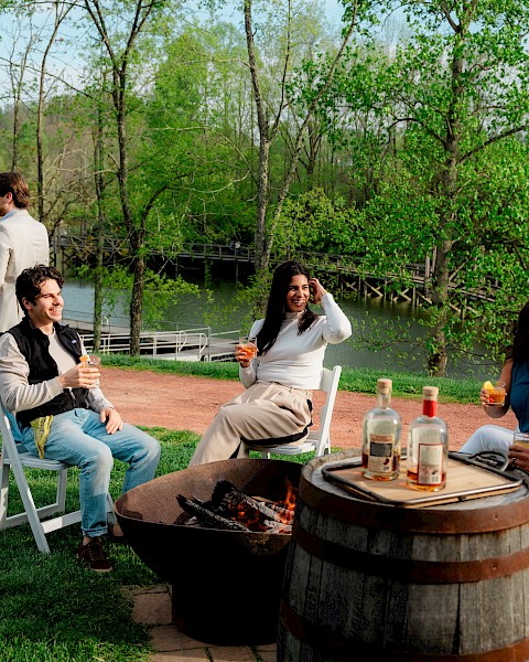 People are enjoying a spring outdoor gathering by a lake, chatting, drinking, and relaxing around a fire pit in a lush, green setting.