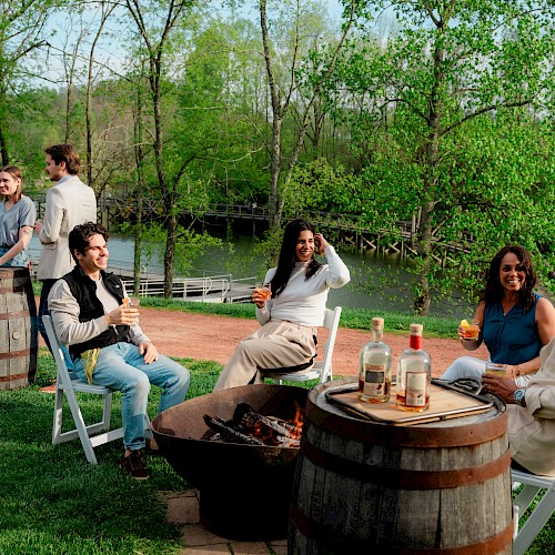 People are enjoying a spring outdoor gathering by a lake, chatting, drinking, and relaxing around a fire pit in a lush, green setting.