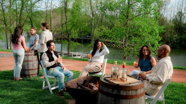 People are enjoying a spring outdoor gathering by a lake, chatting, drinking, and relaxing around a fire pit in a lush, green setting.