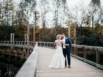 A couple in wedding attire embraces on a wooden bridge surrounded by trees and a body of water in a serene, outdoor setting.