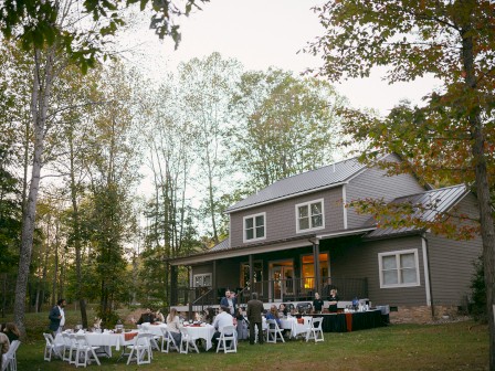 People gather for an outdoor dinner event on a lawn, with a house and trees in the background during the evening.