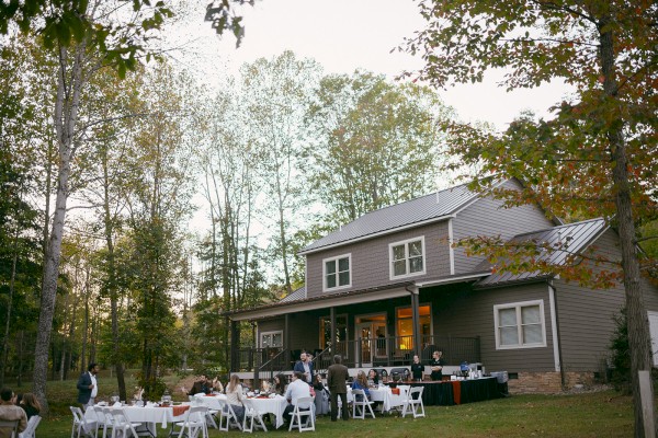 People gather for an outdoor dinner event on a lawn, with a house and trees in the background during the evening.