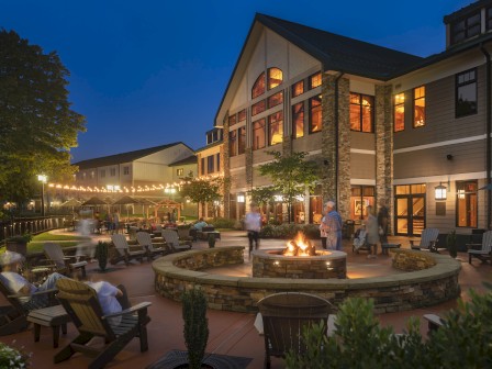 An outdoor seating area with a fire pit, surrounded by people at a large building with string lights and trees in the background during the evening.