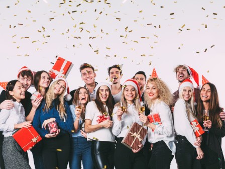 A group of friends celebrating Christmas with gifts, party hats, champagne, and falling confetti, smiling joyfully in festive attire.