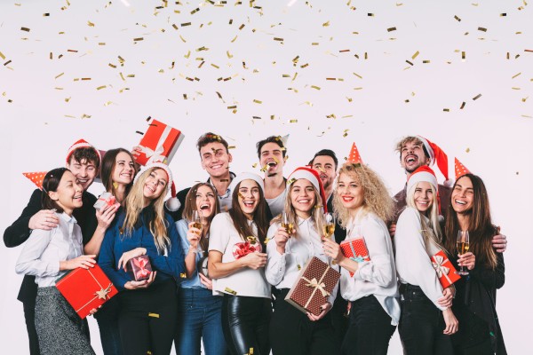 A group of friends celebrating Christmas with gifts, party hats, champagne, and falling confetti, smiling joyfully in festive attire.