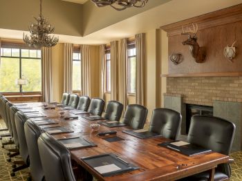 A conference room with a long wooden table, leather chairs, large windows, a fireplace, and deer head decor on the wall ends the sentence.