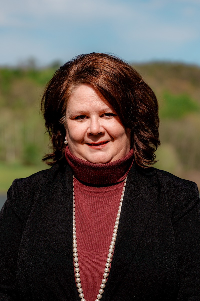 A woman with brown hair wearing a black blazer, a maroon turtleneck, and a pearl necklace outdoors with a blurred green landscape.