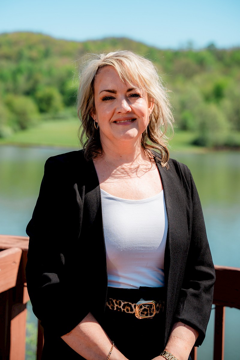 A woman with blonde hair smiles outdoors by a lake with greenery in the background, wearing a black jacket, white top, and leopard print belt.