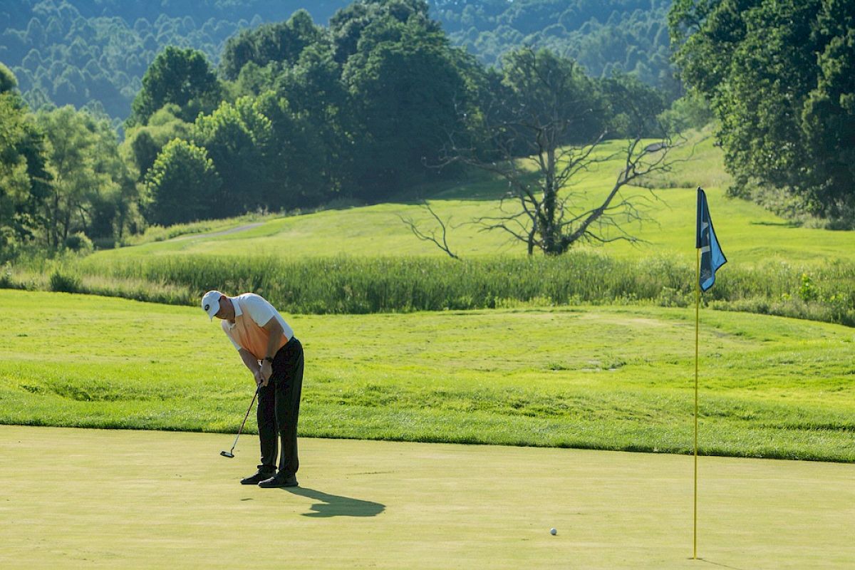 A golfer is putting on a green, with a flagstick nearby on a sunny day, surrounded by lush trees and hills in the background.
