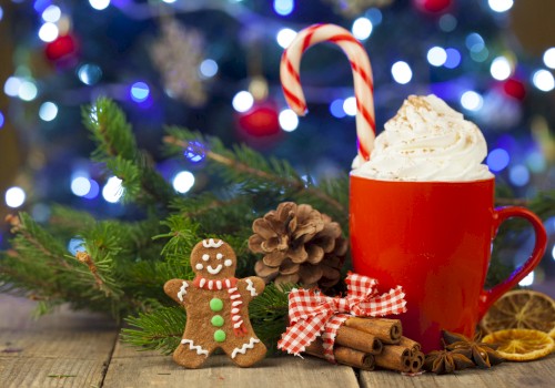 A red mug filled with whipped cream and a candy cane, surrounded by pinecones, cinnamon sticks, dried citrus, and a gingerbread man, set against Christmas lights.