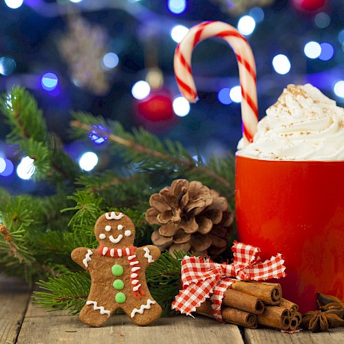 A red mug filled with whipped cream and a candy cane, surrounded by pinecones, cinnamon sticks, dried citrus, and a gingerbread man, set against Christmas lights.