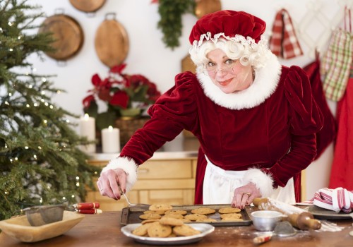 A woman dressed as Santa’s helper is baking cookies in a cozy, decorated kitchen with Christmas decorations and a tree.