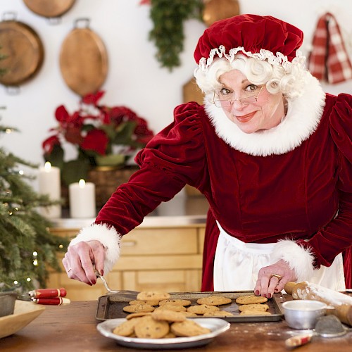 A woman dressed as Santa’s helper is baking cookies in a cozy, decorated kitchen with Christmas decorations and a tree.
