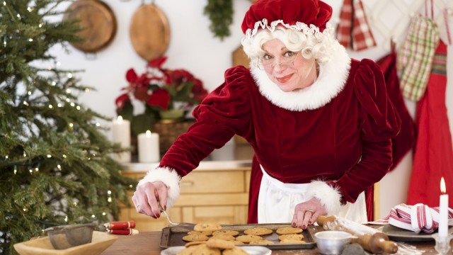A woman dressed as Santa’s helper is baking cookies in a cozy, decorated kitchen with Christmas decorations and a tree.