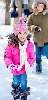 Children are walking through the snow, dressed warmly in winter clothes like jackets, hats, and boots, enjoying a snowy day outdoors.