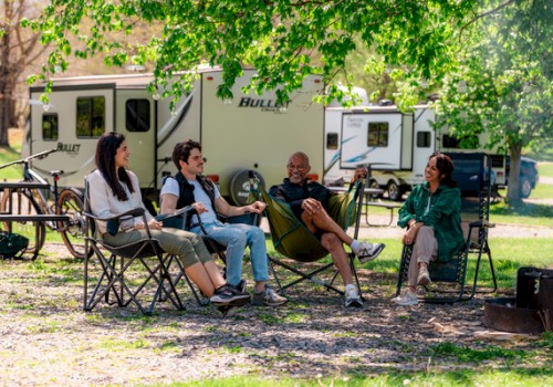 Four people are sitting outdoors near RVs, enjoying a sunny day with trees and camping equipment around them.