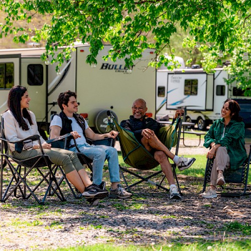 Four people are sitting outdoors near RVs, enjoying a sunny day with trees and camping equipment around them.