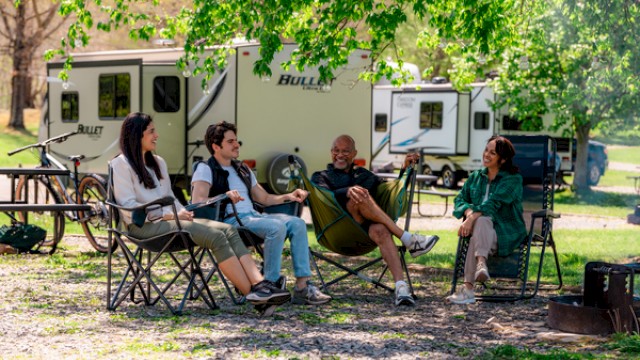 Four people are sitting outdoors near RVs, enjoying a sunny day with trees and camping equipment around them.