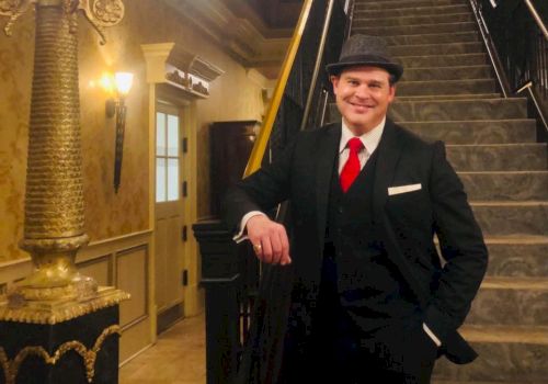 A person in a suit and hat stands in front of an ornate staircase in an elegant interior, leaning against the banister and smiling.