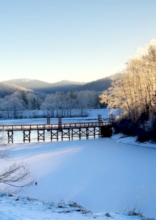 Snowy riverside scene with a wooden bridge, frosty trees, and distant hills bathed in winter sunlight, serene and tranquil.