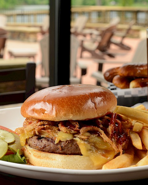A plate with a burger, fries, pickles, and a garnish, alongside a pint of beer, is set on a table with an outdoor seating area in the background.