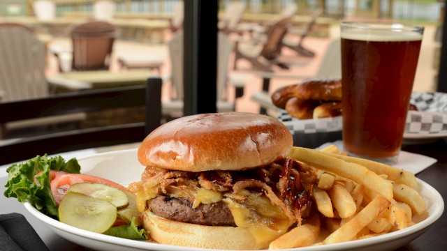 A plate with a burger, fries, pickles, and a garnish, alongside a pint of beer, is set on a table with an outdoor seating area in the background.