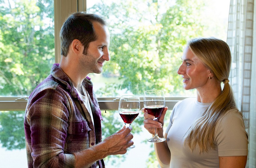 A man and woman enjoy red wine together by a window with lush green trees outside.