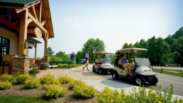 People arriving at a golf course, boarding golf carts near a clubhouse on a sunny day.