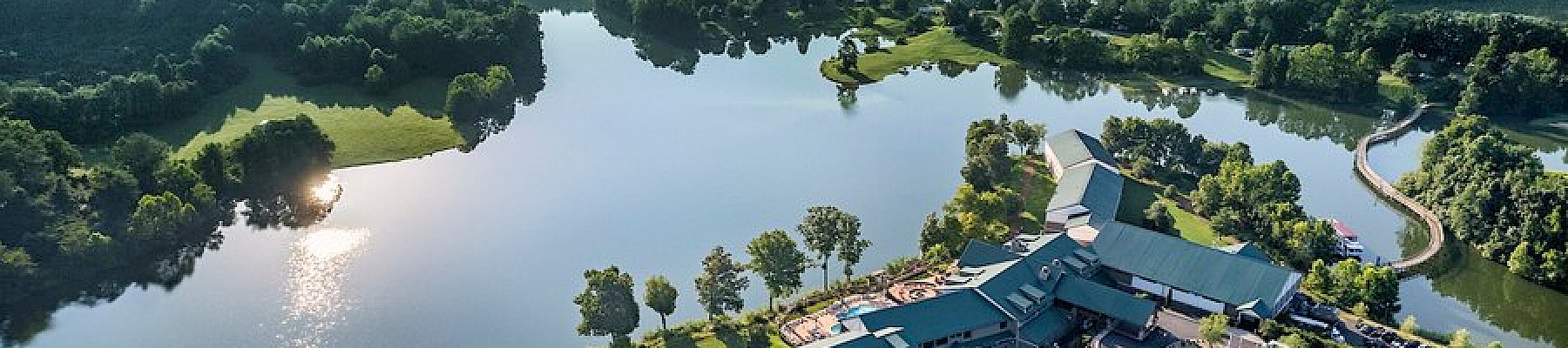 A large building complex with a lake, surrounded by forested mountains and lush greenery, under a clear sky.