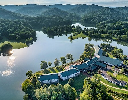 A large building complex with a lake, surrounded by forested mountains and lush greenery, under a clear sky.