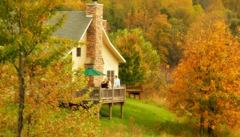 A house with a chimney is surrounded by colorful autumn trees, with people enjoying a conversation on the deck.