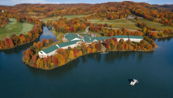 This is an aerial view of a large, white building complex with green roofs, situated on a scenic island surrounded by a calm lake.