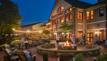 People gather around a lit fire pit outside a large, well-lit modern house during the evening, enjoying a cozy outdoor social setting.