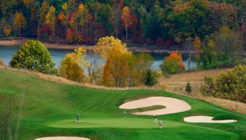 Golfers play on a lush green course near colorful autumn trees and a serene lake in the background of this picturesque scene.