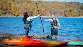 A woman and man laugh and reach out to each other on a lakeshore with colorful autumn trees, paddleboards, and life vests.