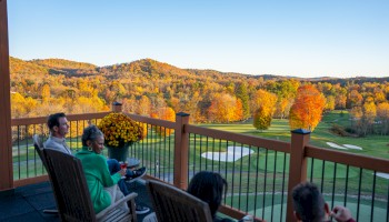 People enjoy a scenic view of a golf course surrounded by autumn-colored trees from a balcony.