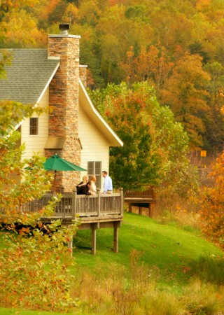 People gather on a house’s deck surrounded by vibrant autumn-colored trees and lush green grass.