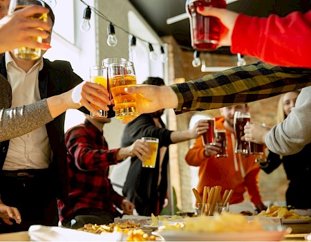 People cheer with drinks at a lively gathering or party, clinking glasses and enjoying food and beverages in a warm indoor setting.