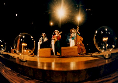 A band performs on stage with old-fashioned instruments, under bright lights, with light bulbs on ground.