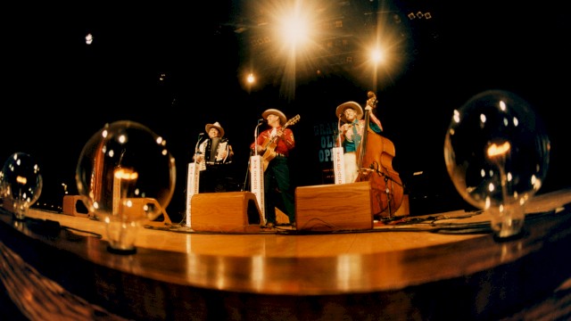A band performs on stage with old-fashioned instruments, under bright lights, with light bulbs on ground.
