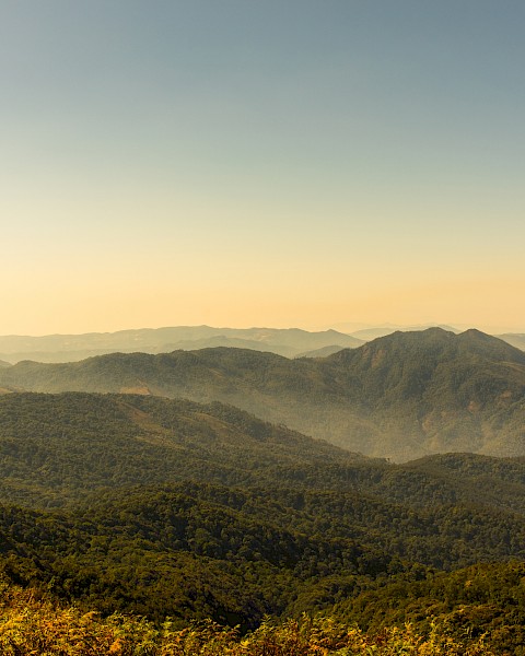 A scenic view of mountain ranges under a clear sky during dusk, showcasing lush greenery and distant rolling hills in a tranquil landscape.