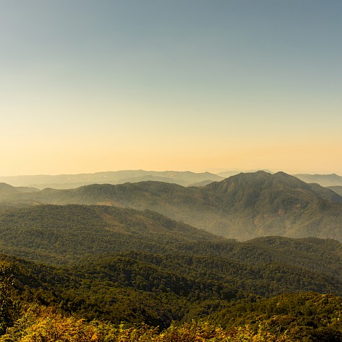 A scenic view of mountain ranges under a clear sky during dusk, showcasing lush greenery and distant rolling hills in a tranquil landscape.