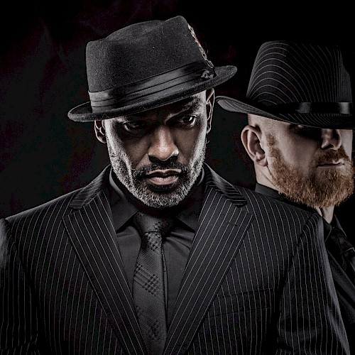 Two men dressed in black suits and hats,pose dramatically against a dark background in a cinematic style.